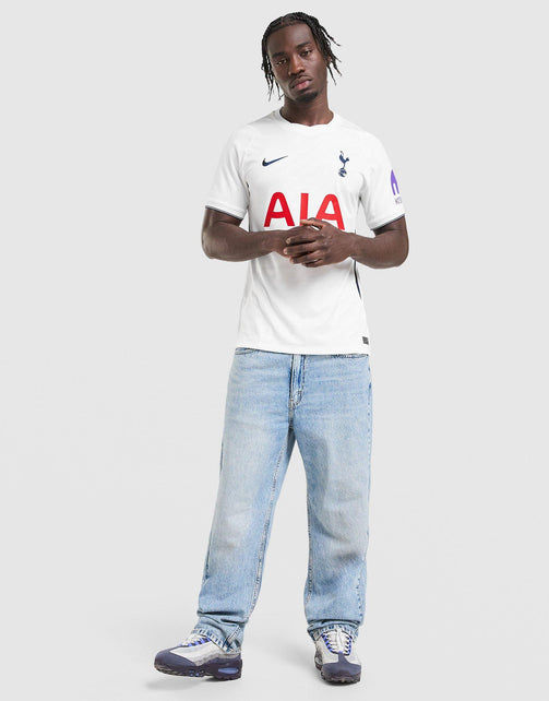 A man modeling a white Nike Tottenham Hotspur 2026/27 Stadium Home Dri-FIT Soccer Jersey with a large red AIA sponsor logo on the front, paired with light blue jeans and sneakers, against a light gray studio background.
