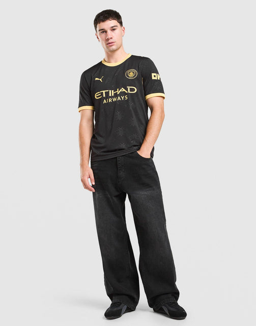 A man modeling a black Manchester City 2026/27 Away Jersey  with gold accents, paired with black jeans and black sneakers, against a light gray studio background.