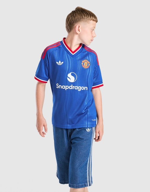 A young boy wearing a blue Adidas Manchester United Junior 26/27 Away Jersey with Snapdragon sponsor logo, paired with blue shorts, photographed against a light gray studio background.