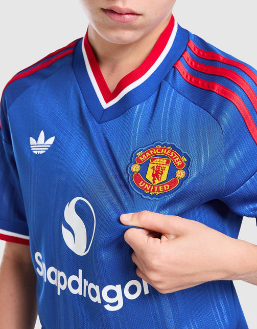 A close-up of a person wearing a blue Manchester United Junior 26/27 Away Jersey with red and white trim, featuring the Adidas logo, Manchester United crest, and sponsor logo.
