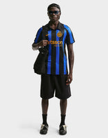 A model wearing a Nike Inter Milan 2026/27 Stadium Home Dri-FIT Soccer Replica Jersey with blue and black vertical stripes, black shorts, and black sneakers, carrying a black bag against a light gray background.