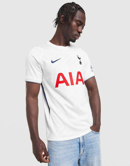 A man wearing a white Nike Tottenham Hotspur 2026/27 Stadium Home Dri-FIT Soccer Jersey with red AIA sponsor text, blue swoosh and cockerel logos, paired with light blue jeans, against a light gray background.