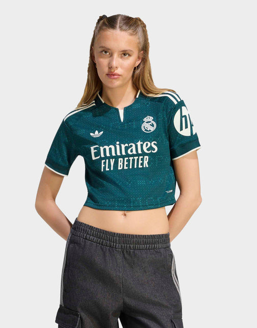 A young woman modeling a dark teal Real Madrid crop top soccer jersey with white accents, featuring the Adidas logo, Real Madrid crest, and Emirates Fly Better sponsor text, paired with dark gray cargo shorts against a light gray background.