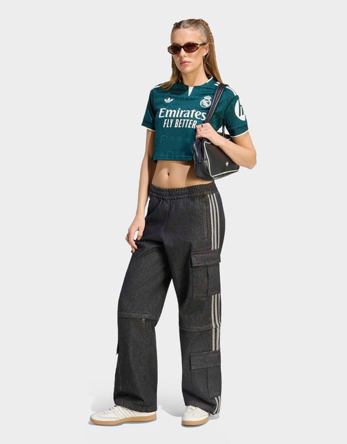 A model wearing a dark green Real Madrid cropped t-shirt with Emirates Fly Better branding, paired with dark gray wide-leg cargo pants featuring white side stripes, white sneakers, and a black shoulder bag, set against a light gray background.