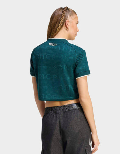 A woman shown from the back wearing a Real Madrid 26/27 Cropped Fan Jersey dark teal cropped short-sleeve polo shirt with a subtle tonal pattern and white trim, paired with dark gray pants, against a light gray studio background.