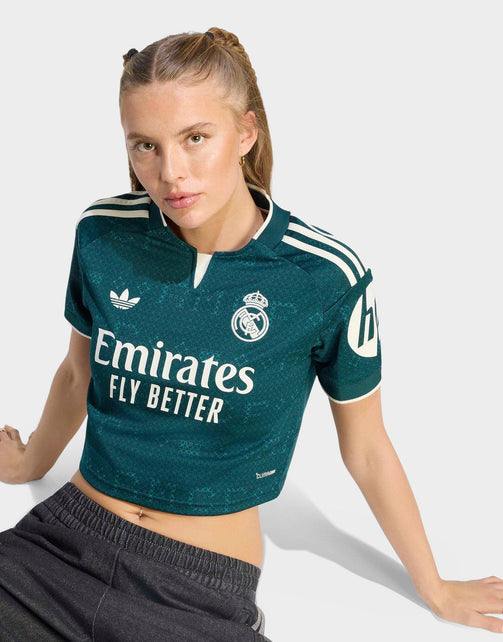 A woman modeling a dark teal Real Madrid 26/27 Cropped Fan Jersey with white sponsor and brand logos, paired with dark gray sweatpants against a light gray studio background.
