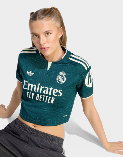 A woman modeling a dark teal Real Madrid 26/27 Cropped Fan Jersey with white sponsor and brand logos, paired with dark gray sweatpants against a light gray studio background.