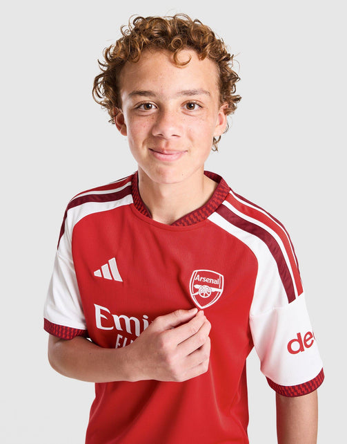 A young boy with curly brown hair wearing an adidas Arsenal FC 26/27 Home Jersey Junior's, featuring a red and white design with the club's crest on the chest, photographed against a light gray background.