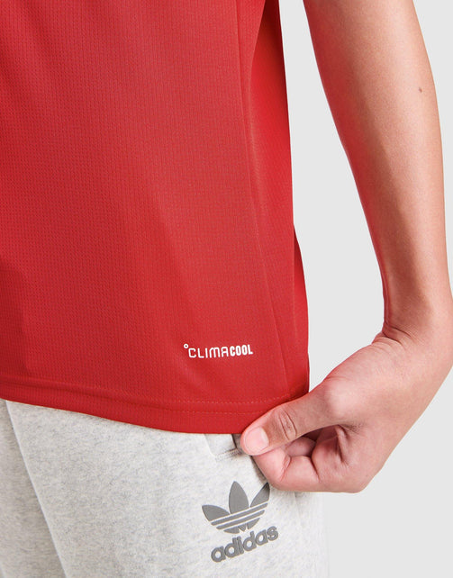 Close-up of a person wearing a adidas Arsenal FC 26/27 Home Jersey Junior's climacool athletic top and light gray Adidas sweatpants, with the hand adjusting the hem of the top.