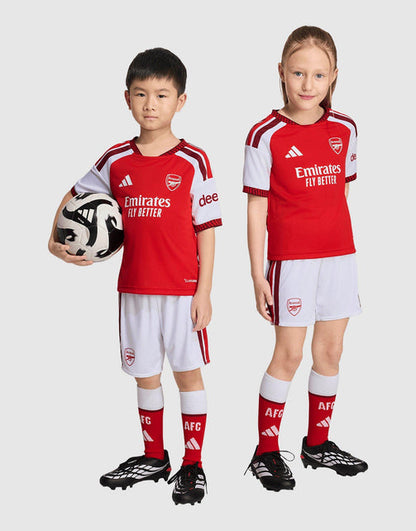 Two children modeling matching red and white adidas Arsenal FC 26/27 Home Set Children's soccer kits, including jerseys, shorts, socks, and cleats, against a light gray studio background. One child holds a soccer ball.