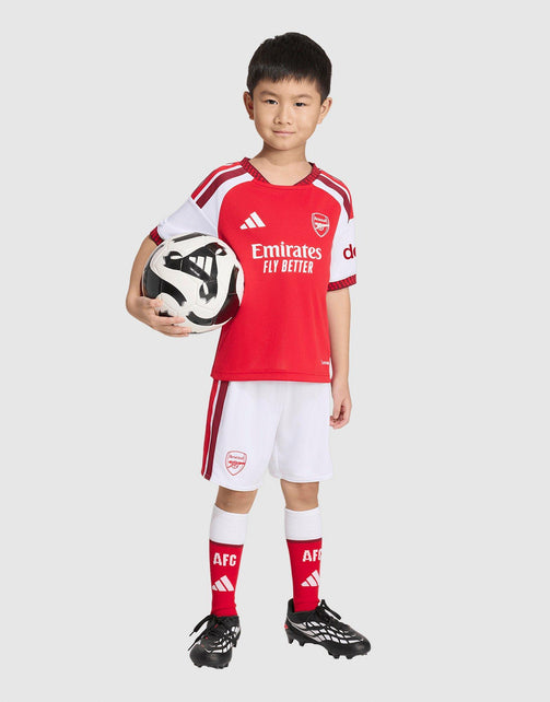 A young boy modeling an adidas Arsenal FC 26/27 Home Set Children's soccer kit, featuring a red and white jersey with 'Emirates Fly Better' sponsor logo, white shorts with red stripes, red socks, and black soccer cleats, while holding a black and white soccer ball against a light gray background.