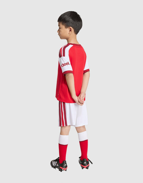 A young boy modeling a red and white adidas Arsenal FC 26/27 Home Set Children's soccer kit, featuring a red jersey with white shoulder stripes and 'Diesel' branding, white shorts with red stripes, red socks, and black cleats, set against a solid light gray background.