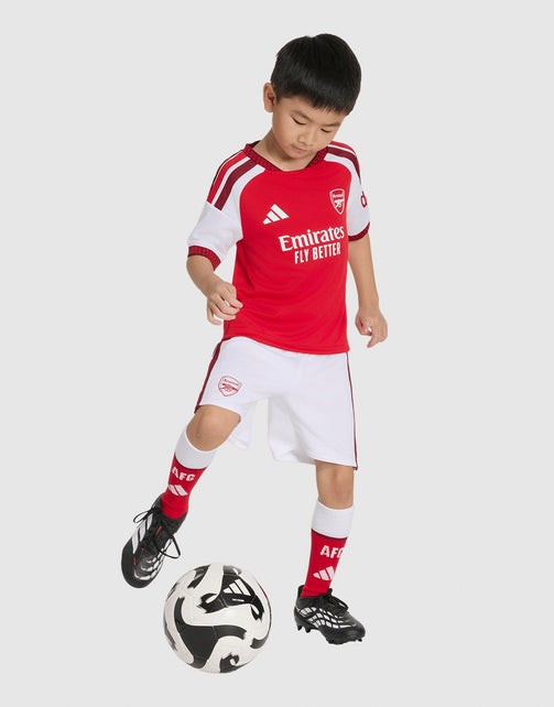 A young boy wearing a red and white adidas Arsenal FC 26/27 Home Set Children's soccer kit with matching shorts, socks, and cleats, dribbling a black and white soccer ball against a light gray background.