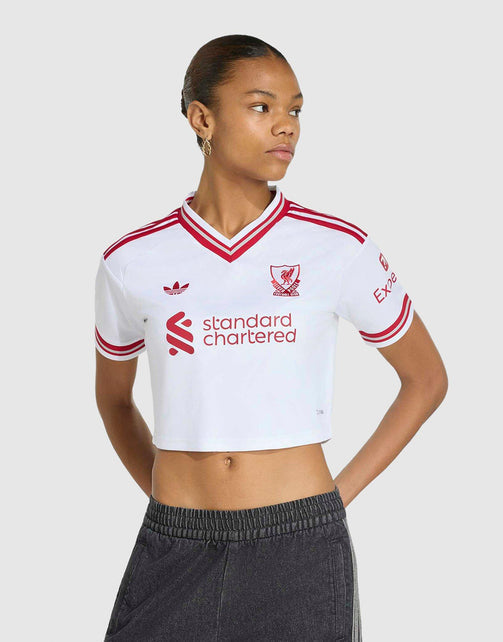 A model wearing aadidas Liverpool FC 26/27 Away Jersey Crop with red stripes and logos, featuring the Adidas logo and Liverpool crest, paired with dark gray sweatpants against a light gray background.