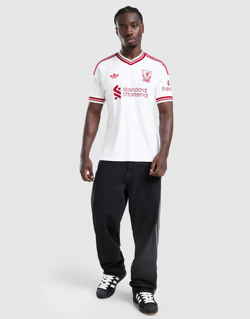 A man modeling a white adidas Liverpool FC 26/27 Away Jersey with red trim, paired with black pants and black-and-white sneakers, against a light gray studio background.