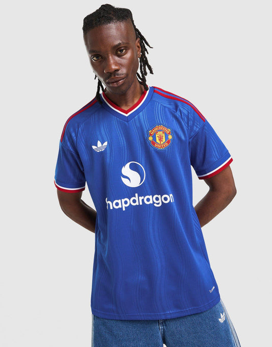 A man wearing a blue adidas Manchester United 26/27 Away Jersey with red and white trim, featuring the Adidas logo, Manchester United crest, and Shapdragon sponsor logo, set against a plain light gray background.