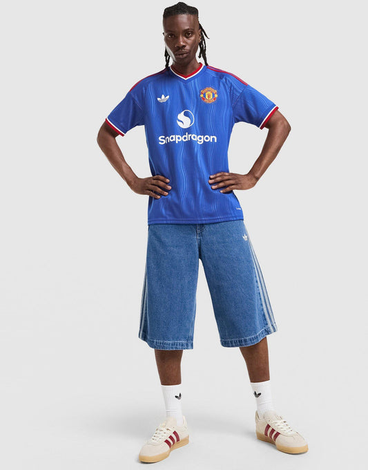 A model stands against a light gray studio background wearing a blue adidas Manchester United 26/27 Away Jersey with red and white trim, blue denim shorts, white socks, and cream Adidas sneakers. Visible branding includes the Manchester United crest, Adidas logo, and sponsor text 'Spadragon'.
